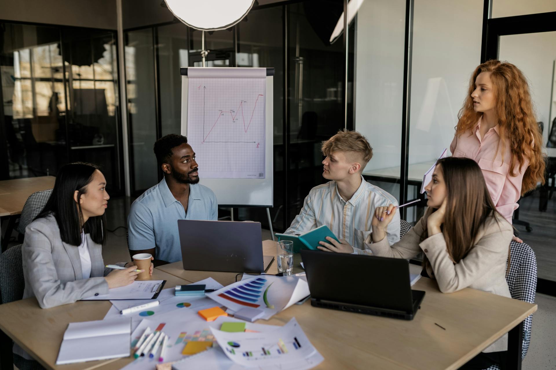 Diverse team of professionals discussing data analytics in a modern glass office with laptops and whiteboard charts